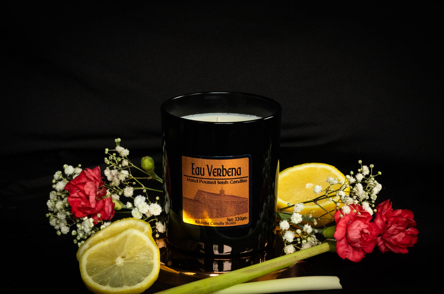 Copper Soy Wax Black Jar candle with 'Eau Verbena' branding surrounded by lemons and flowers and lemongrass on a dark background