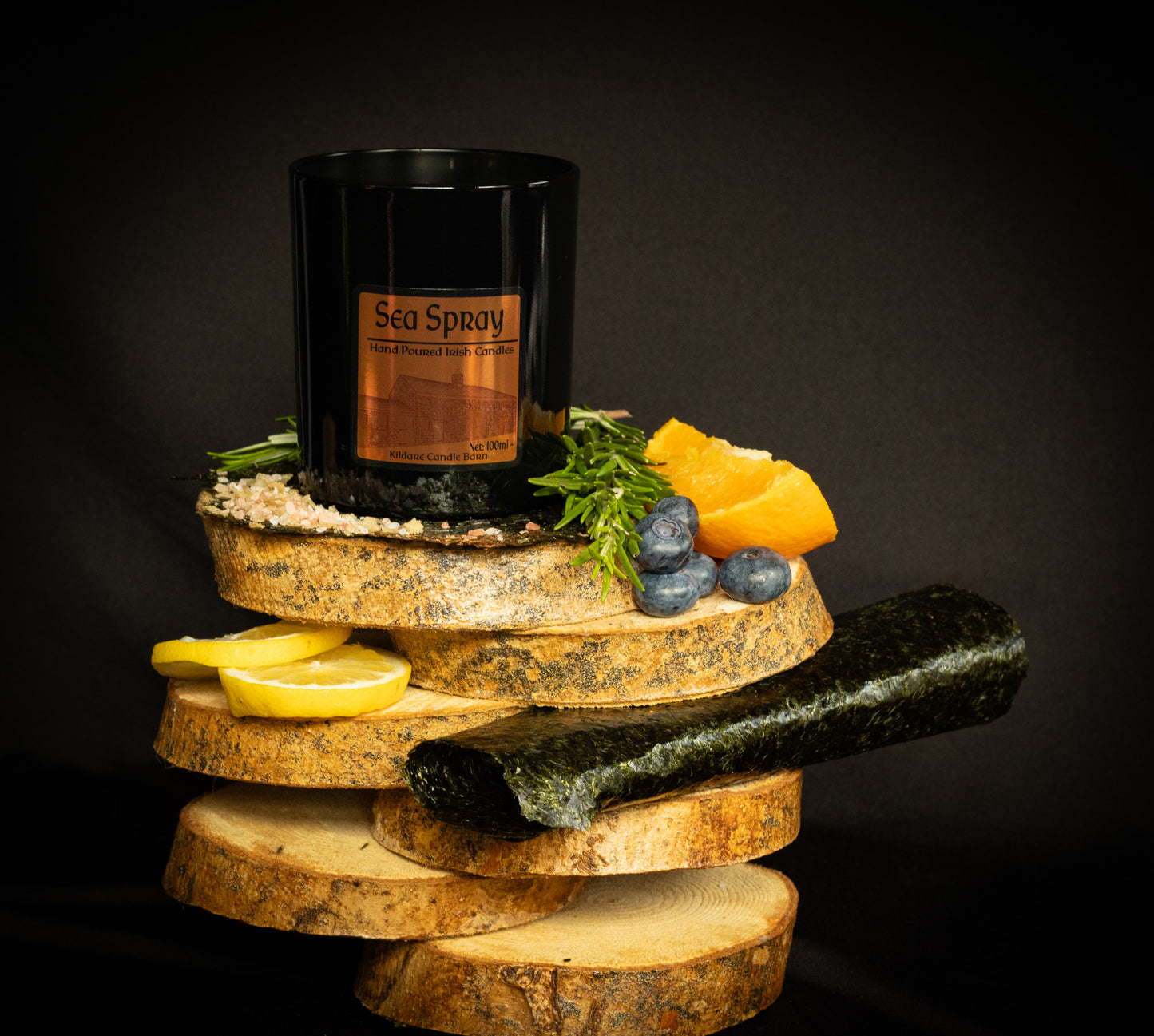 Irish Soy Wax Candle labeled 'Sea Spray'. Black Jar Candle on a wooden display with lemons, stones, and herbs & seaweed against a dark background