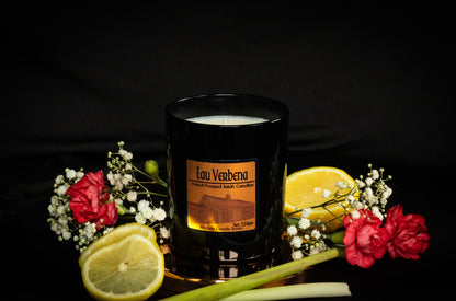 Copper Soy Wax Black Jar candle with 'Eau Verbena' branding surrounded by lemons and flowers and lemongrass on a dark background