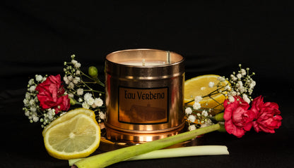 Copper Soy Wax tin candle with 'Eau Verbena' branding surrounded by lemons and flowers and lemongrass on a dark background