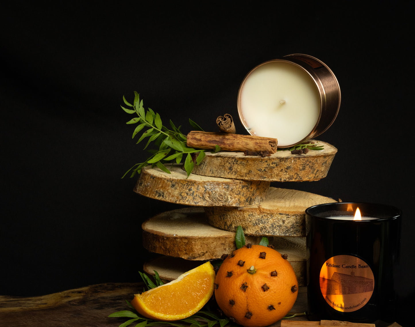 Irish Soy Wax Candle in a tin with a wooden coaster, orange slice, cinnamon stick, and clove studded clementine on a dark background.